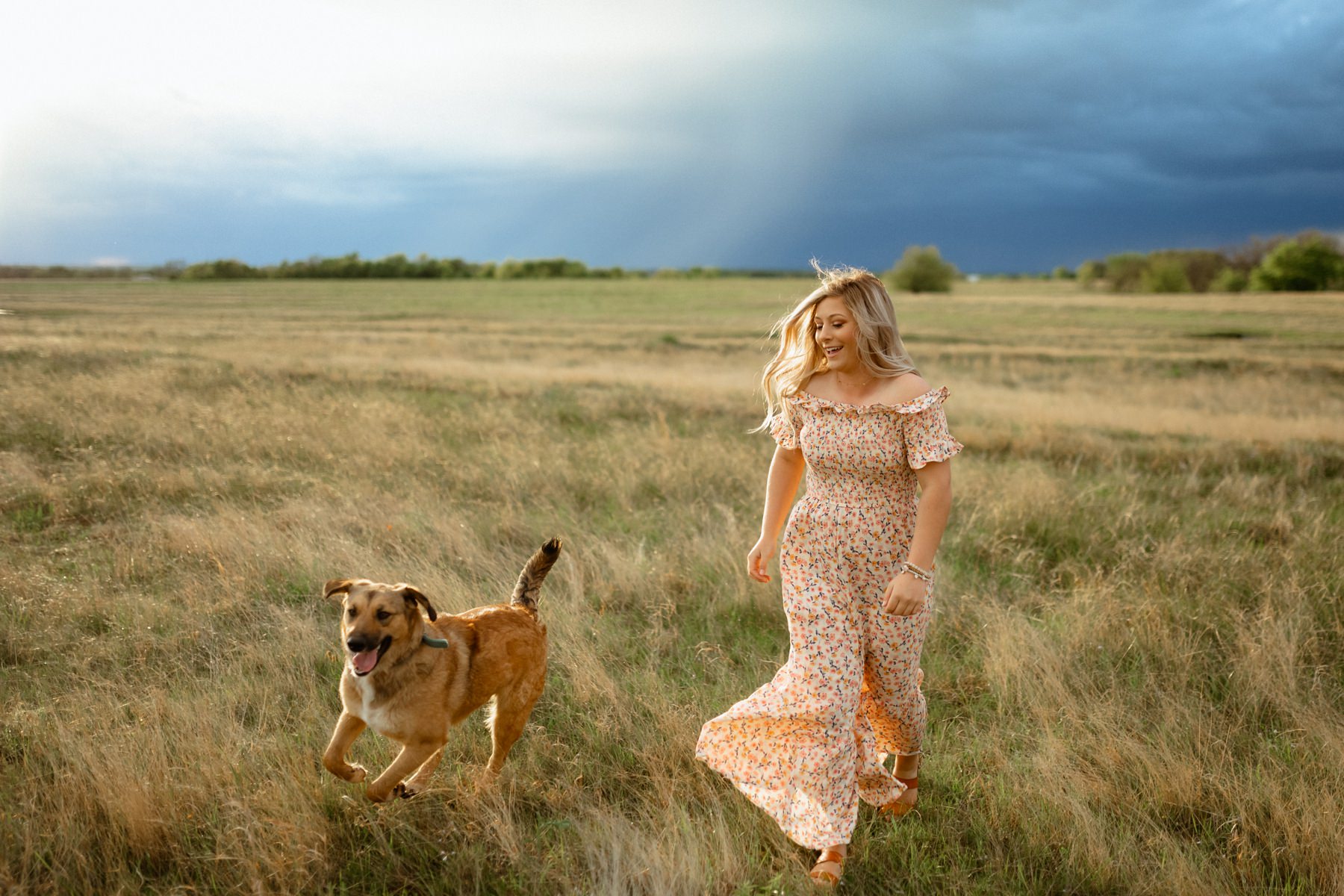 A girl in a floral romper running with her golden labrador in an open field at her senior photo session.